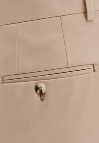 Beżowa tkanina o gładkiej strukturze, z tylną kieszenią i zapięciem na złoty guzik. Subtelne detale szwów wokół kieszeni.