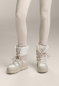 Beżowe zimowe buty z wyściółką, o fakturowanej powierzchni, z przodu na sznurowadła i grubą podeszwą. Noszone na przylegających legginsach.