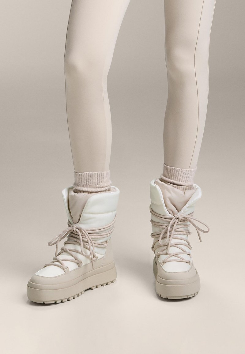Beżowe zimowe buty z wyściółką, o fakturowanej powierzchni, z przodu na sznurowadła i grubą podeszwą. Noszone na przylegających legginsach.