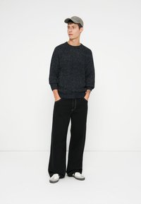 Mörk texturerad tröja med rund halsringning, matchad med vida svarta byxor och vita sneakers. En grå keps fulländar outfiten.