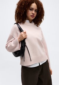 Γυναίκα με σγουρά μαλλιά που φοράει ανοιχτό ροζ πουλόβερ πάνω από λευκό πουκάμισο, σκούρο παντελόνι και κρατάει μια μαύρη τσάντα ώμου.