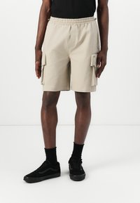Beige cargo shorts with an elastic waistband, two side pockets and two large side cargo pockets, paired with black sneakers and socks.