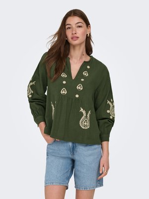 Moteris dėvi žalią ilgomis rankovėmis megztinį su smėlio spalvos siuvinėjimu ir šviesiai mėlynas džinsines šortus, stovinti viena ranka kišenėje priešais vientisą foną.