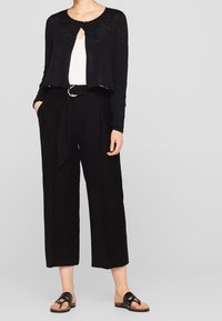 Svart kort cardigan med långa ärmar, knappstängning, lager på en vit topp. Svarta högmidjade vida byxor med knytbälte och svarta sandaler.