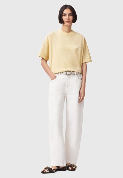 Personne aux cheveux courts et foncés portant un t-shirt ample jaune pâle, un pantalon blanc à jambes larges, une ceinture beige et des ballerines noires à motifs, debout devant un fond uni.