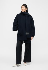 Γυναίκα που φοράει ένα φαρδύ ναυτικό παλτό με ένα ταιριαστό κασκόλ, μαύρο ευρύχωρο παντελόνι και λευκές μυτερές γόβες, στέκεται μπροστά σε ένα απλό φόντο.