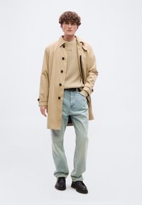 Jeune homme debout portant un trench-coat beige par-dessus un pull marron clair, un jean bleu clair et des chaussures noires devant un fond uni clair.