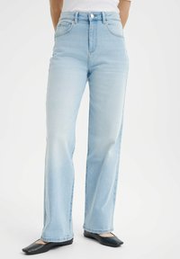 Jeans de tiro alto en un azul claro, confeccionados en tela de mezclilla y con un diseño de pierna ancha. Presentan costuras visibles y bolsillos delanteros, complementados con zapatos planos negros.