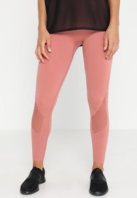Žena v růžových bezešvých legínách s texturovanými kolenními panely a černými skládacími sportovními botami, stojící na bílém pozadí.