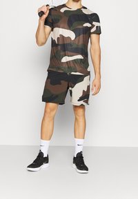 Koszulka z krótkim rękawem w kamuflażowym wzorze oraz szorty w brązowych, zielonych i kremowych kolorach, w połączeniu z czarnymi butami sportowymi i białymi skarpetkami.