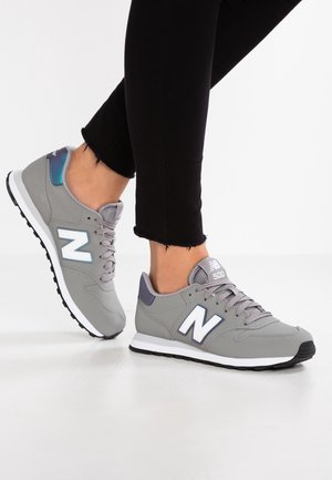 Jambes en pantalon noir portant des baskets New Balance 500 grises avec des semelles blanches, debout sur une surface blanche.