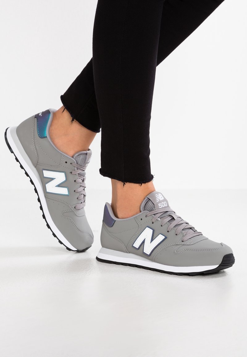 Jambes en pantalon noir portant des baskets New Balance 500 grises avec des semelles blanches, debout sur une surface blanche.
