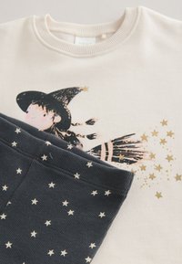 Sweat-shirt crème avec un motif de sorcière, des étoiles dorées, et un pantalon bleu foncé avec un motif d'étoiles blanches. Les deux articles sont en tissu doux et de coupe décontractée.