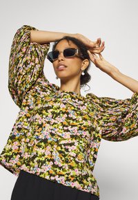 Top com estampa floral em tons de verde, rosa e azul; apresenta mangas bufantes e um decote em forma de barco; combinado com óculos de sol retangulares pretos.