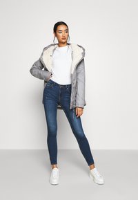 Grå polstret jakke med faux fur-fôr, kombinert med en hvit ribbet topp, blå skinny jeans og hvite joggesko. Jakken har hette og lommer.