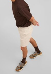 Hnědá košile s krátkým rukávem, béžové šortky a hnědé a šedé tenisky s bílou podrážkou. Ponožky jsou tmavě hnědé. Jednoduchý design, volný střih.