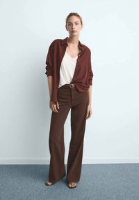 Pantalon large marron et chemise boutonnée bordeaux sur un haut blanc ; tissu lisse, coupe décontractée, avec col ouvert et manches retroussées.