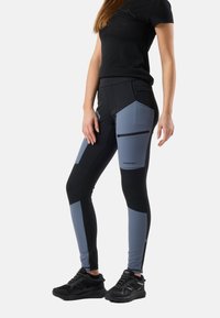 Kvinde iført sorte og grå tætsiddende bukser med lynlåslomme på låret og sorte sneakers, stående mod en hvid baggrund.