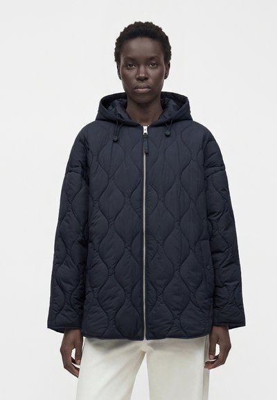 Personne portant une veste matelassée à capuche bleu marine avec une fermeture éclair à l’avant et un pantalon blanc, debout devant un fond clair uni.