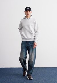 Sudadera gris con bolsillo frontal, jeans de mezclilla azul, gorra negra y zapatillas grises. El atuendo presenta un estilo casual con un ajuste relajado.