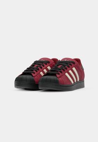 Red suede sneakers with black laces and toe cap, featuring three white stripes on the side and a black rubber sole. Round shape design.