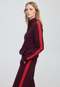 Jeune femme en survêtement bordeaux avec des rayures rouges sur les manches et les côtés, se tenant avec les mains dans les poches, faisant légèrement face sur le côté.
