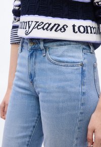 Κοντινό πλάνο ατόμου που φοράει ανοιχτό μπλε τζιν και μπλε πουλόβερ με λευκή λωρίδα που αναγράφει το κείμενο "tommy jeans".