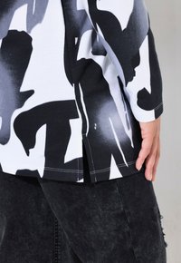 Maglietta a maniche lunghe in bianco e nero con un audace motivo astratto, dotata di spacchi laterali e una texture morbida visibile nel tessuto.