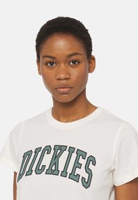 Weißes Baumwoll-T-Shirt mit kurzen Ärmeln, auf dem ein großes grünes und schwarzes "DICKIES"-Logo über die Brust gedruckt ist. Glatte Textur, klassischer Rundhalsausschnitt.