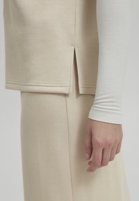 Top beige a maniche corte con fessura laterale, realizzato in tessuto morbido e liscio. Lo strato sottostante ha maniche lunghe di un beige più chiaro.