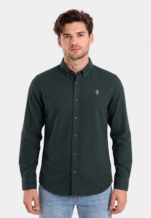 Joven con camisa de botones verde oscuro con un pequeño logo blanco de árbol en el pecho y vaqueros azul claro, de pie frente a un fondo blanco.
