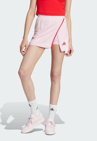 Svetle růžová sportovní sukně s postranním rozparkem, vyrobená z měkkého materiálu. Obsahuje červený akcent a logo Adidas. Doplňena růžovými botami a bílými ponožkami.