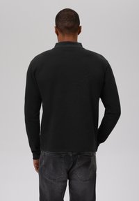 Svart langermet polo skjorte, i bomullsmateriale, med klassisk krage, synlige sømmer, ensfarget, brukt sammen med mørk grå slitte jeans.