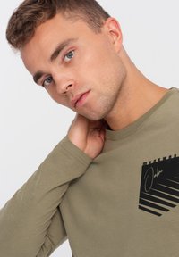 Olivenfarbener Langarm-T-Shirt mit einem schwarzen grafischen Taschendesign mit Streifen. Weicher Stoff und lockere Passform mit rundem Ausschnitt.