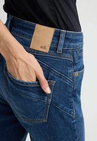 Los jeans de mezclilla azul oscuro presentan un parche de cuero marrón con la etiqueta "adl", costuras visibles y una mano colocada en el bolsillo trasero.