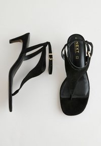 Zwarte hoge hakken sandalen met een dunne enkelband, bovenwerk van suede en een glanzende zwarte voetbed met gouden logo-details.