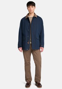 Chaqueta de tela azul marino con cuello de pana marrón, botones a presión, dos bolsillos frontales y cierre de cremallera, llevada sobre un suéter gris.