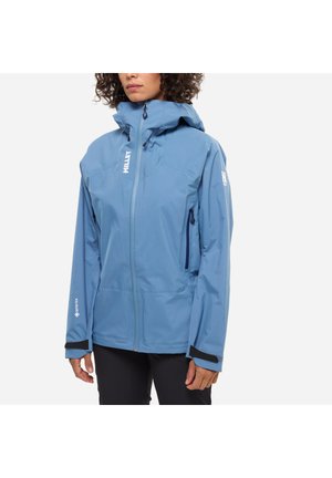 Femme portant une veste imperméable bleu clair avec capuche, fermeture éclair avant, poches latérales zippées et poignets ajustables sur fond blanc.