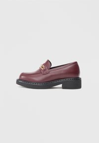 Burgundy läderloafer med svart kraftig sula, med en guldfärgad metallkedja över vaden och slät textur.