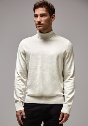 Homme portant un pull col roulé beige clair et un pantalon noir, se tenant contre un fond gris uni, regardant sur le côté.