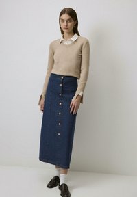 Lichtbruin v-hals trui over een witte blouse; gecombineerd met een lange denimrok met roségouden knopen aan de voorkant.