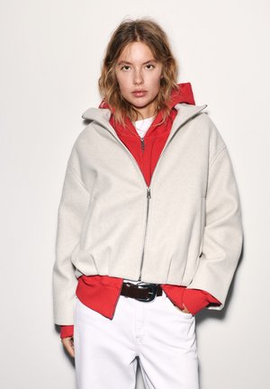 Jeune femme aux cheveux lâches et ondulés portant un sweat à capuche rouge zippé sous une veste zippée gris clair et un pantalon blanc avec une ceinture noire.