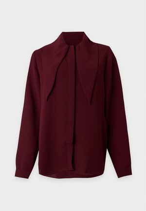 Femme aux longs cheveux raides portant un chemisier ample bordeaux avec un grand col et des sandales noires à talons hauts avec des brides à la cheville.