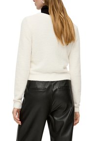 Weißer strukturierten Pullover mit langen Ärmeln, kombiniert mit schwarzen Lederhosen. Der Pullover hat einen taillierten Saum und einen hohen Kragen.