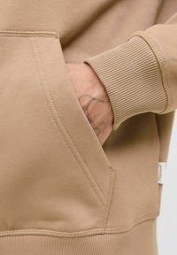 Beżowa bluza z kangurzą kieszenią, ściągaczami w mankietach i miękką strukturą materiału. Widoczne detale szycia podkreślają design.