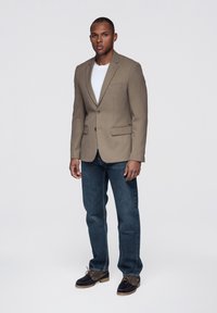 Beżowa, dopasowana marynarka z klapami z wycięciami, zapinana na jeden guzik, z dwoma przednimi kieszeniami, zestawiona z niebieskimi dżinsami i brązowymi butami.