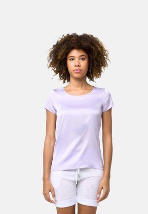 Femme aux cheveux bouclés portant un t-shirt à manches courtes violet clair et un short blanc transparent, debout devant un fond uni.
