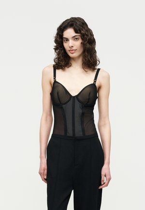 Keharapärasega naine kannab musta läbipaistvast korsetikujulist ülaosa reguleeritavate rihmadega ja kõrge vöökohaga musti pükse ühtlase tausta ees.