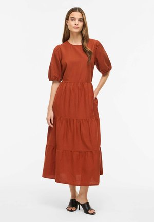 Kobieta w rdzawym, warstwowym midi z bufkami i czarnych, otwartych sandałach na obcasie, stojąca z jedną ręką w kieszeni.