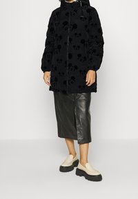 Chaqueta de plumas negra con patrones en relieve de Mickey Mouse, cierre de cremallera y capucha; combinada con una falda de cuero negra y zapatillas crema sin cordones.